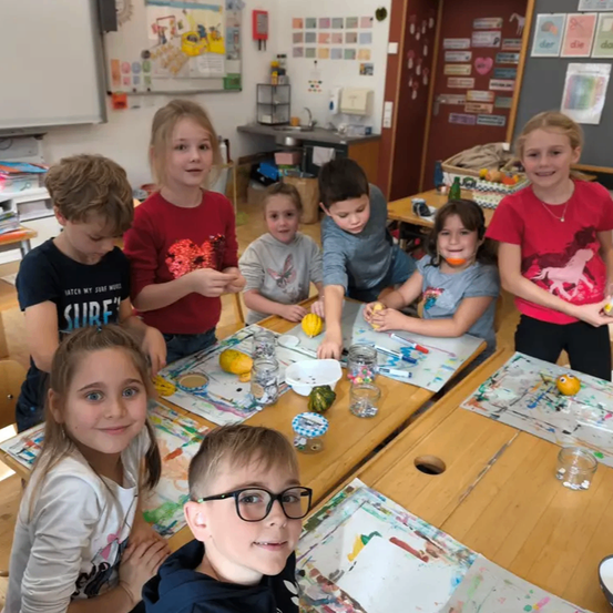 Eine Gruppe von Kindern versammelt sich um einen Tisch in einem Klassenzimmer und ist in einer Kunstaktivität engagiert. Sie haben verschiedene Kunstmaterialien vor sich, und einige tragen Brillen. Der Raum ist gut dekoriert mit farbenfrohen Postern an der Wand.
