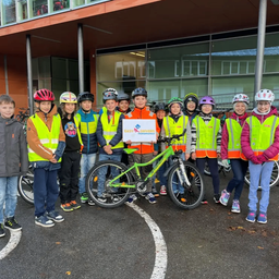 Eine Gruppe von Kindern in Sicherheitswesten und Helmen posiert für ein Foto mit einem grünen Fahrrad. Sie stehen vor einem modernen Gebäude mit großen Fenstern.