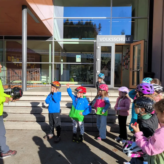 Eine Gruppe von Kindern mit Helmen versammelt sich vor einem Gebäude mit einer Glastür, auf der Volksschule steht. Ein Mann in einem gelben Mantel steht links.