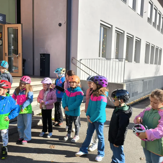Eine Gruppe von Kindern mit Helmen und Jacken steht vor einem weißen Gebäude mit Glasfenstern und einer Treppe.