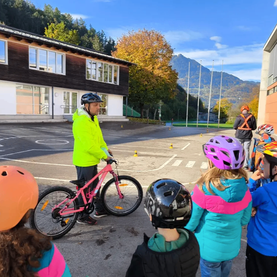 Ein Mann in einer gelben Jacke bringt Kindern das Fahrradfahren bei. Sie tragen alle Helme und stehen um den Mann und das Fahrrad herum.