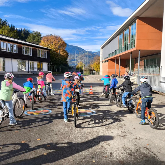 Eine Gruppe von Kindern in Helmen fährt auf einer Straße mit Gebäuden und Bergen im Hintergrund mit Fahrrädern.