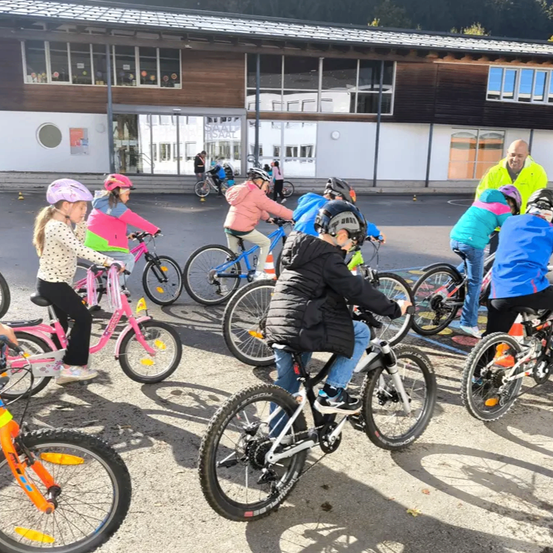Kinder fahren mit erwachsener Aufsicht auf einem Schulparkplatz mit Fahrrädern. Einige tragen Helme und andere Jacken. Hinter ihnen steht ein Gebäude mit Glasfenstern.