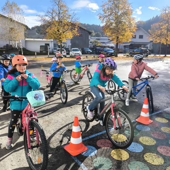 Kinder in Helmen fahren auf einem gepflasterten Bereich mit farbigen Kreisen mit Fahrrädern, umgeben von geparkten Autos und Gebäuden.