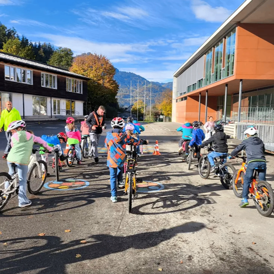 Mehrere Kinder fahren auf einem asphaltierten Weg vor einem Gebäude mit Fahrrädern. Einige Erwachsene beaufsichtigen.