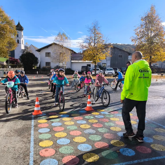 Eine Gruppe von Kindern fährt auf einem Parkplatz mit Fahrrädern, wobei ein Mann in einer neongrünen Jacke in der Nähe steht. Bunte Kreise sind auf dem Boden gemalt.