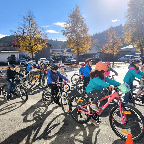 Eine Gruppe junger Radfahrer steht in einer Reihe, alle tragen Helme, auf einer gepflasterten Fläche. Bäume und Gebäude sind im Hintergrund zu sehen.