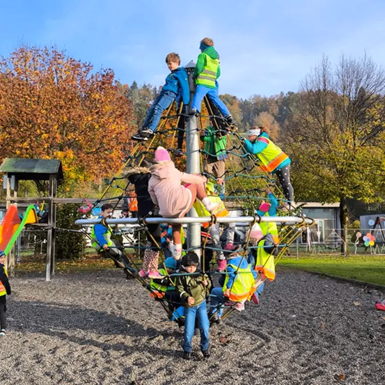 Kinder spielen auf einer großen Kletterstruktur in einem Outdoor-Spielplatz an einem sonnigen Tag. Einige klettern, während andere am Fuß stehen.