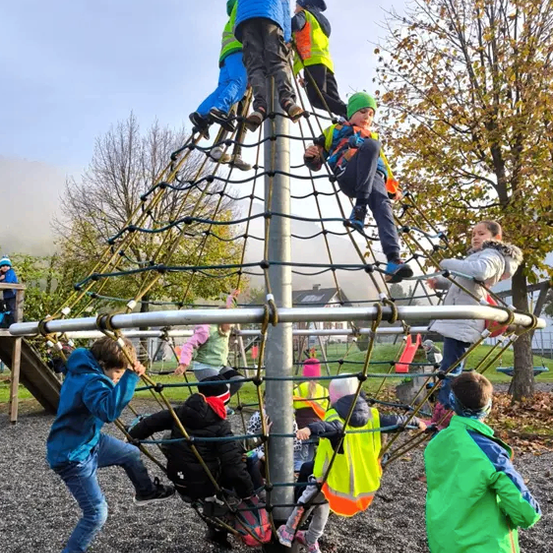 Kinder klettern in einem großen Netzgerüst im Spielplatz. Einige tragen Sicherheitswesten. Bäume und Häuser sind im Hintergrund.