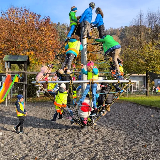 Eine Gruppe von Kindern klettert und spielt auf einer großen kugelförmigen Struktur in einem Outdoor-Spielplatz, umgeben von Bäumen und einem Gebäude.