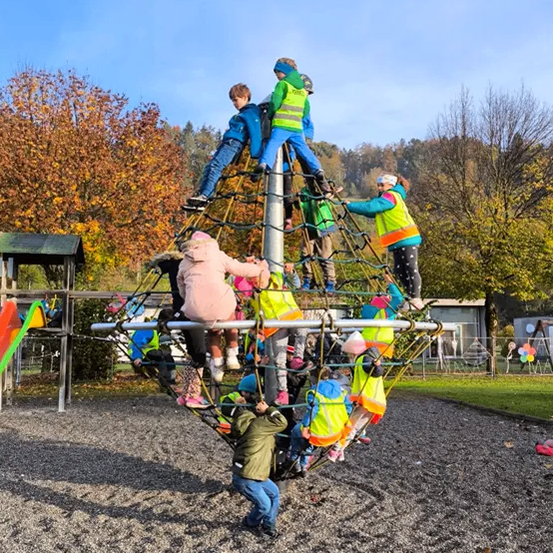 Mehrere Kinder klettern in einem Park auf einer großen Netzstruktur. Einige tragen Sicherheitswesten. Bäume und ein Gebäude sind im Hintergrund.