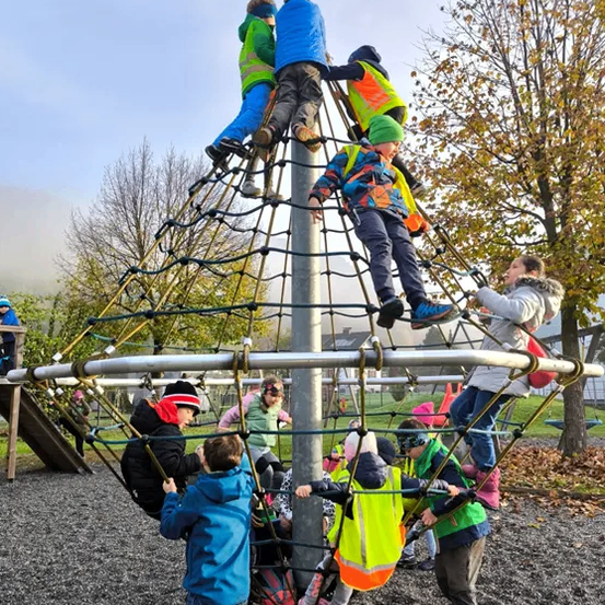 Mehrere Kinder spielen auf einem Klettergerüst im Freien, tragen Westen und Helme. Bäume und ein Gebäude sind im Hintergrund.