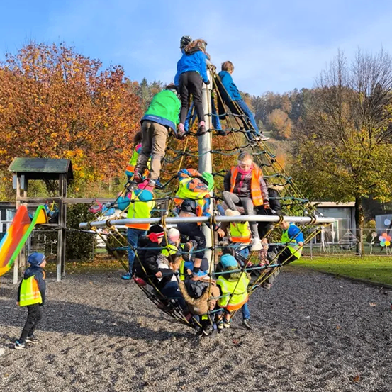 Kinder spielen auf einer kugelförmigen Kletterstruktur in einem Spielplatz. Mehrere Kinder klettern und andere sitzen. Ein Gebäude und Bäume sind im Hintergrund.