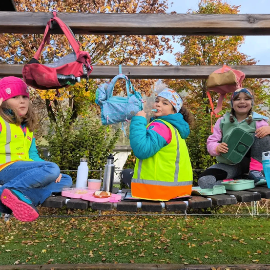 Drei junge Mädchen in Sicherheitswesten sitzen auf einer Holzbank im Freien, eine putzt sich die Zähne, eine andere isst und die dritte lächelt. Verschiedene Taschen und Flaschen hängen über ihnen.