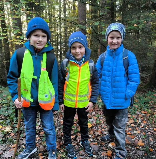 Drei junge Jungen stehen in einem Wald. Einer hält einen Stock, und alle tragen Sicherheitswesten. Sie tragen Winterkleidung und scheinen zu lächeln.