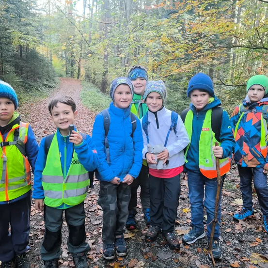 Eine Gruppe von Kindern, gekleidet in reflektierende Westen und Mützen, steht in einem Wald und lächelt für ein Foto. Sie sind von Bäumen und gefallenen Blättern umgeben.
