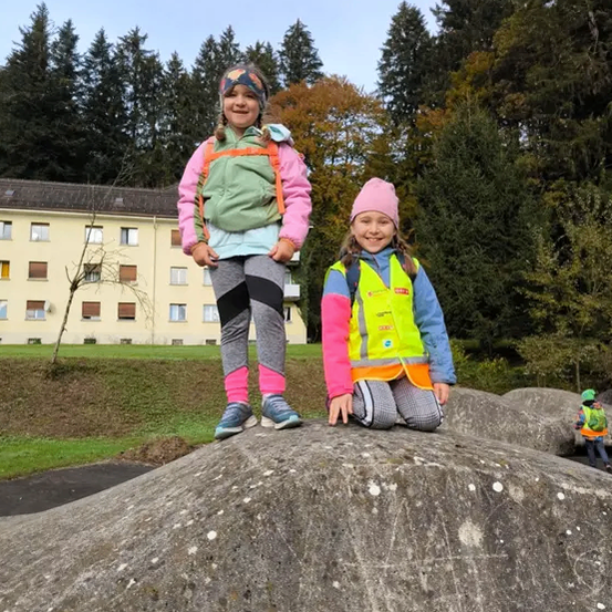 Zwei junge Mädchen stehen auf einem großen Felsen in einem Park. Hinter ihnen sind Bäume und ein Gebäude. Ein Mädchen trägt eine grüne Weste und das andere ein rosa Jackett.
