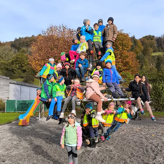 Eine Gruppe von Kindern in Jacken und Mützen posiert für ein Foto auf einem Spielplatz. Einige sitzen auf einer Struktur, während andere stehen. Bäume und ein Gebäude sind im Hintergrund zu sehen.