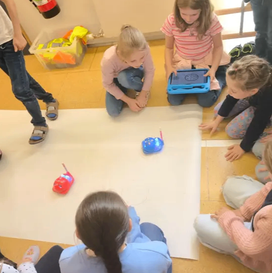 Mehrere Kinder sitzen auf dem Boden um ein weißes Blatt herum und steuern Spielzeugautos mit Tablets. Ein Feuerlöscher ist an der Wand montiert.