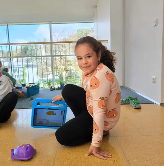 Ein junges Mädchen kniet auf einem gefliesten Boden und hält ein Tablet in den Händen. Sie lächelt und schaut auf das Tablet, das ein farbenfrohes Diagramm anzeigt. Sie trägt ein langärmeliges Hemd mit Kürbis-Motiv. Ein lila Spielzeugauto steht in der Nähe. Der Raum hat Glasfenster mit Blick auf Bäume und Gebäude.