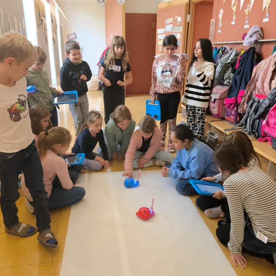 Mehrere Kinder sind um einen Roboter auf dem Boden versammelt und halten Tablets. Eines Mädchen steht in der Nähe eines Kleiderständers.