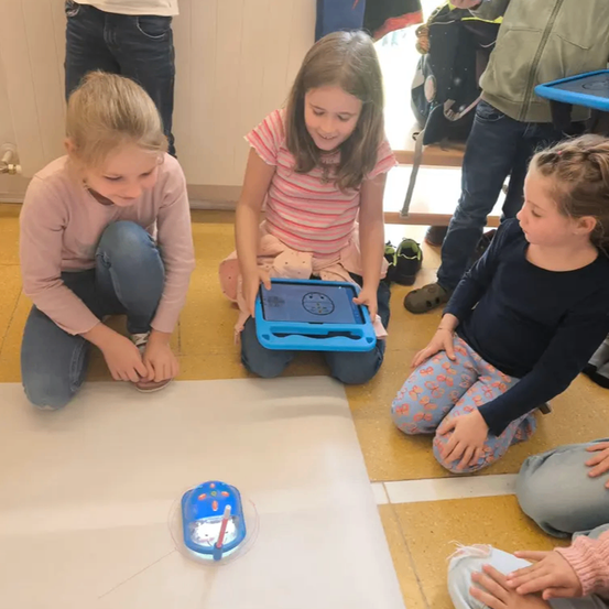 Drei Mädchen sitzen auf dem Boden, zwei konzentrieren sich auf ein Tablet und eines schaut einen kleinen Roboter an. Sie sind in einem Raum mit gelben Fliesen.
