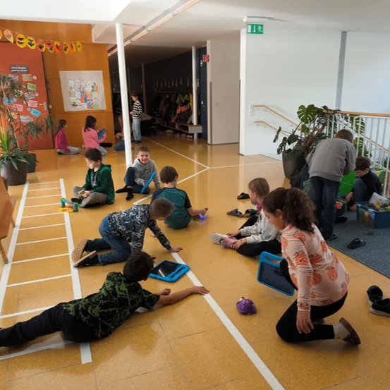 Kinder in einem Schulraum sitzen und liegen auf dem Boden, benutzen Tablets und Spielzeug. Einige tragen Turnschuhe. Eine Frau kniet mit einem Tablet.