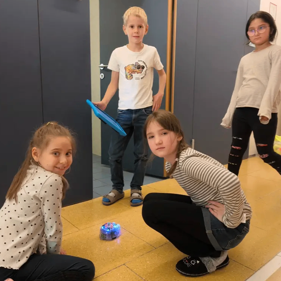 Drei Kinder spielen mit einem Roboter auf dem Boden. Ein Junge hält ein Tablet.