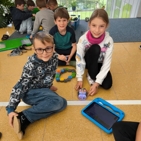 Kinder sitzen auf dem Boden und spielen mit Spielzeug. Ein Junge benutzt ein Tablet, während ein Mädchen einen Roboter hält. Andere beschäftigen sich mit Spielzeug auf dem Boden.