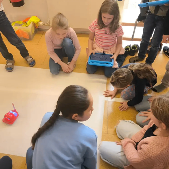 Mehrere Kinder sitzen auf dem Boden und beschäftigen sich mit einem Tablet. Ein Mädchen kniet und hält ein Tablet, während ein anderes Mädchen mit einem roten Spielzeugauto in der Nähe aufmerksam zuschaut.