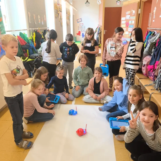 Eine Gruppe von Kindern in einem Klassenzimmer spielt mit Robotern auf einem weißen Boden. Sie halten Tablets und einige stehen in der Nähe der Wand.