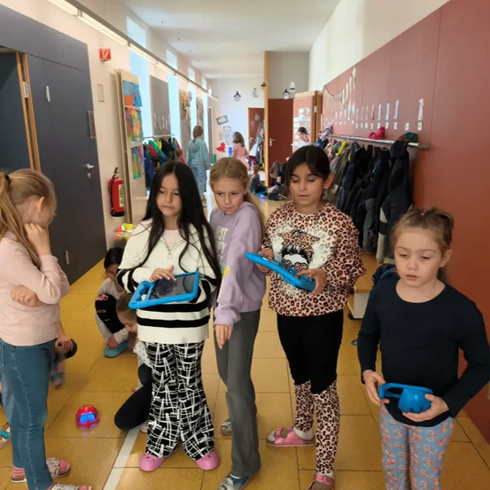 Fünf Mädchen stehen in einem Flur. Sie alle haben Tablets in den Händen und schauen darauf. Der Flur hat geflieste Böden und braune Wände mit Kleidern, die an Gestellen hängen.