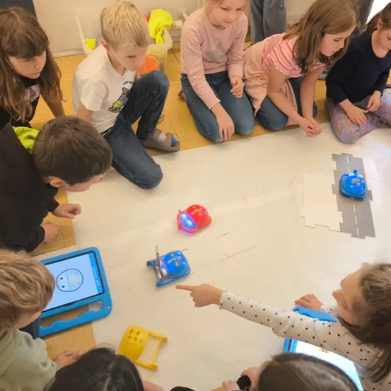 Mehrere Kinder sitzen im Kreis um eine große Tafel und betrachten kleine Roboter auf dem Boden. Vor ihnen stehen einige Tablets.