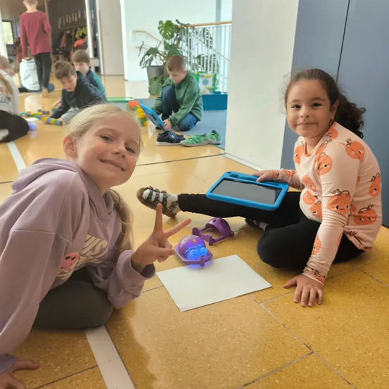 Zwei junge Mädchen sitzen auf dem Boden mit Spielzeug um sie herum. Eine hält ein Tablet, während die andere einen kleinen Roboter hat.