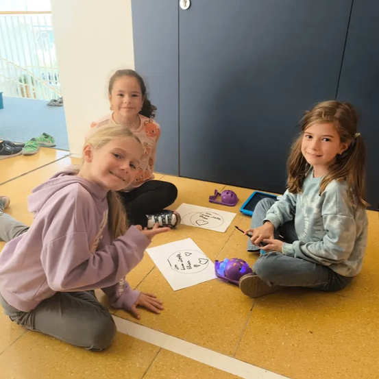 Drei Mädchen sitzen auf dem Boden in einem Klassenzimmer. Sie sind an einer Aktivität beteiligt, die Zeichnungen und einen Roboter beinhaltet. Ein Mädchen lächelt, während ein anderes den Roboter betrachtet.