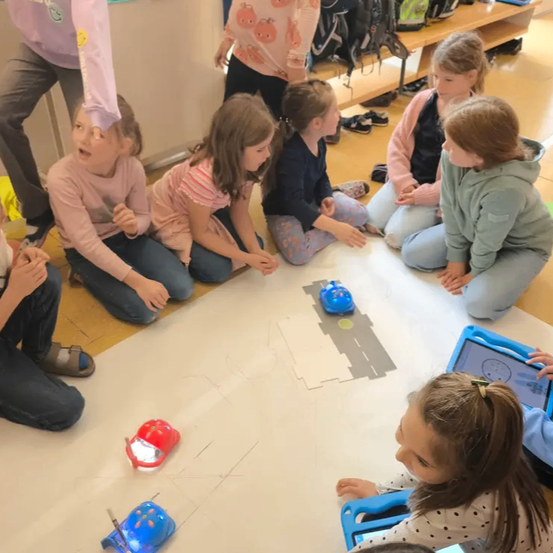 Eine Gruppe von Kindern sitzt im Kreis auf dem Klassenzimmerboden. Sie haben zwei Roboter vor sich auf dem Boden. Ein Kind schaut in die Kamera.
