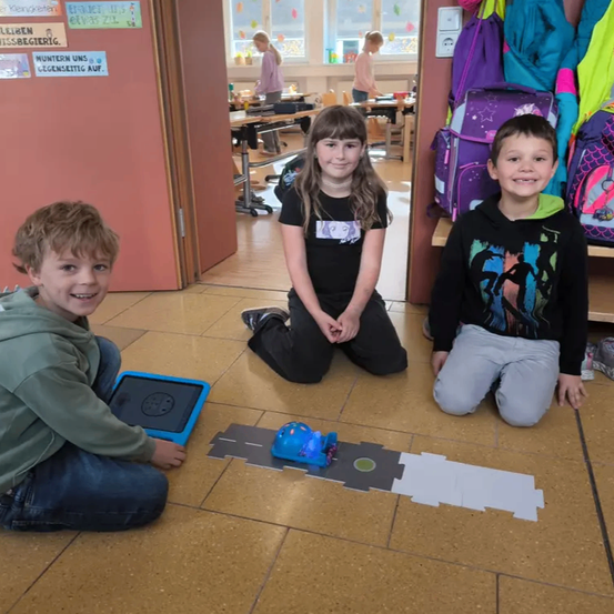 Drei Kinder sitzen auf dem Boden in einem Klassenzimmer und sind in einer Aktivität vertieft. Ein Junge hält ein Tablet, während ein Mädchen neben ihm sitzt. Ein weiterer Junge sitzt neben ihr. Sie sind umgeben von einem Spielzeugauto auf einer Rennstrecke und bunten Postern an der Wand.