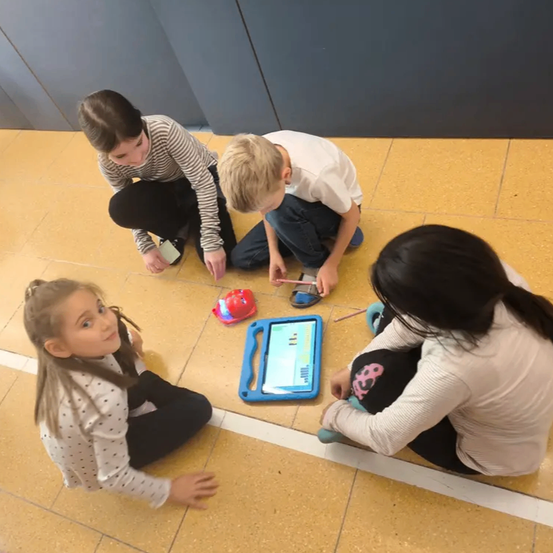 Vier Kinder sitzen auf dem Boden und sind in einer Aktivität vertieft. Drei konzentrieren sich auf ein Tablet, während das vierte ein kleines rotes Objekt hält. Sie befinden sich in einem Klassenzimmer mit gelbem Boden.