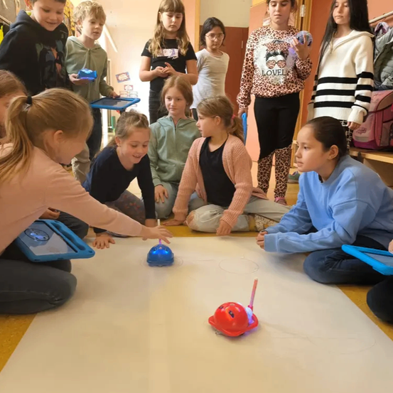 Eine Gruppe von Kindern in einem Klassenzimmer beobachtet einen Roboter auf einem weißen Blatt Papier, wobei einige Tablets verwenden und andere hinter ihnen stehen.
