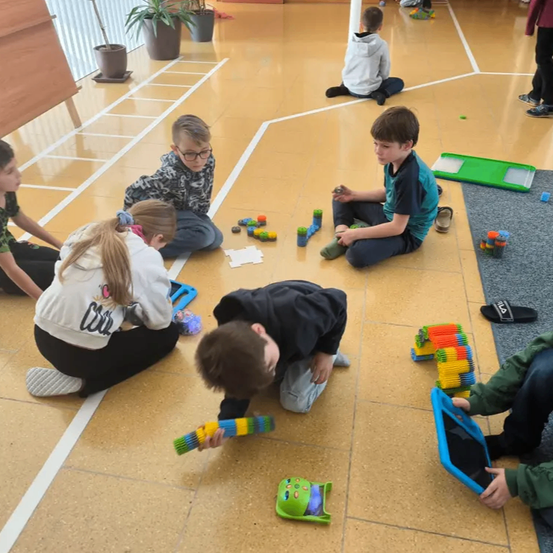 Mehrere Kinder spielen mit bunten Bauklötzen auf einem gefliesten Boden. Einige sitzen auf dem Boden, während andere knien. Eine grüne Ablage mit weiteren Bauklötzen ist in der Nähe.