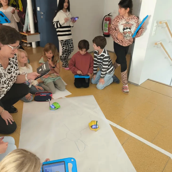 Kinder sitzen mit Tablets auf dem Boden. Eine Frau sitzt links und hält ein Tablet. Zwei Kinder stehen mit einem Tablet und einem Paddel. Ein Feuerlöscher ist an der Wand montiert.