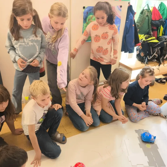 Eine Gruppe von Kindern ist um eine große Papierzeichnung auf dem Boden versammelt, einige kniend und andere stehend. Sie scheinen sich auf ein blaues Spielzeugauto auf dem Papier zu konzentrieren.