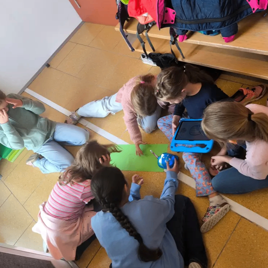 Eine Gruppe von Kindern sitzt auf dem Boden. Sie arbeiten an einem grünen Blatt Papier und scheinen ein Kunstprojekt zu machen. Ein Kind hält ein Tablet.