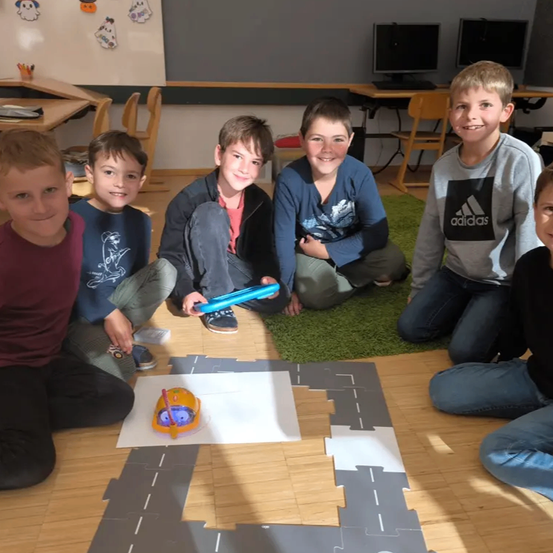 Fünf Jungen sitzen mit einem Spielzeugauto auf einem Puzzle auf dem Boden in einem Klassenzimmer. Ein Junge hält eine Fernbedienung.