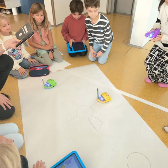 Eine Gruppe von Kindern in einem Klassenzimmer sitzt um ein großes weißes Papier auf dem Boden herum. Sie verwenden Tablets, um Spielzeugroboter zu steuern, die auf das Papier zeichnen. Einige Kinder sitzen auf dem Boden, während andere knien.