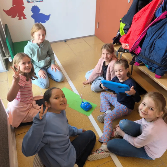 Eine Gruppe von Mädchen sitzt auf dem Boden in einem Raum. Einige lächeln und posieren für ein Foto. Eine grüne Matte liegt auf dem Boden und ein paar Jacken sind auf dem Regal. Ein Kind hält eine Spielzeugpistole in der Hand.