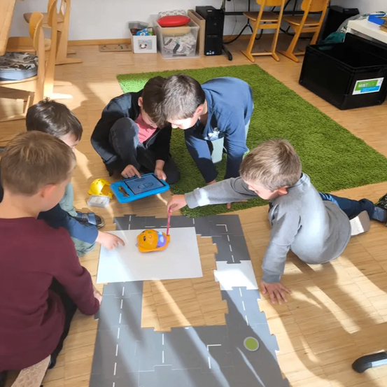 Fünf junge Jungen sitzen auf dem Boden und beschäftigen sich mit einer praktischen Aktivität mit einem Spielzeugauto auf einer Papiermatte. Sie konzentrieren sich auf die Aufgabe, wobei zwei Jungen das Auto steuern und andere genau zuschauen.