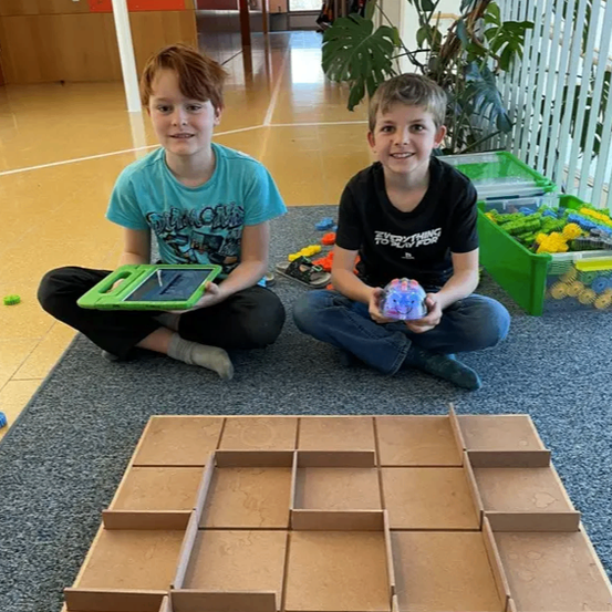 Zwei Jungen sitzen auf dem Boden, einer hält ein Tablet, der andere ein kleines Spielzeug. Vor ihnen steht eine große Pappstruktur mit mehreren Fächern.