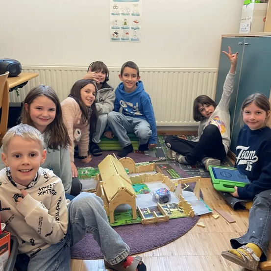 Eine Gruppe von Kindern sitzt um eine hausähnliche Struktur aus Holz auf einem Teppich herum und lächelt für die Kamera. Dahinter befinden sich ein weißer Heizkörper und Poster an der Wand.