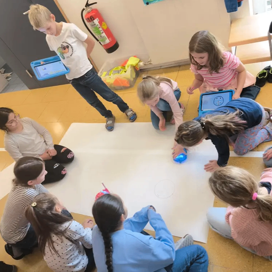 Mehrere Kinder sitzen im Kreis auf dem Boden. Ein Junge steht und hält ein Tablet, um einen Roboter zu steuern. Die Kinder beobachten und nehmen an der Aktivität teil, wobei ein Feuerlöscher im Hintergrund zu sehen ist.